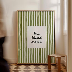 May include: A framed art print with a green and white striped background and the text "How blessed are we." The artwork is displayed in a room with a small wooden stool and a checkered floor. The frame is a light brown color.