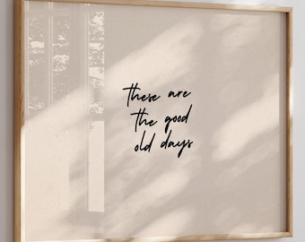 May include: A framed print with the text "these are the good old days" in black script. The print has a beige background and is encased in a light brown wooden frame. The image is displayed on a white wall.