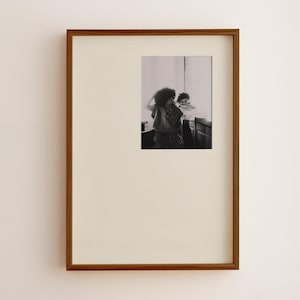 May include: A framed black and white photograph. The photograph depicts two people, one with curly hair, standing near a window. The frame is a dark brown color, and the matting is off-white. The photograph is centered within the frame.