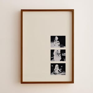 May include: A rectangular wall art piece featuring a dark brown wooden frame and a cream-coloured mat. The centre contains three stacked black and white photographs of a person wearing a towel on their head and a robe, captured in various poses.