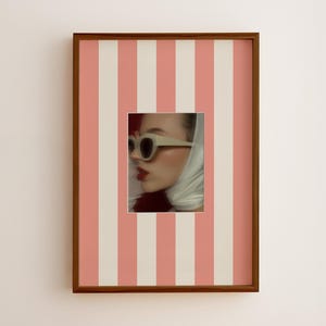 Peut inclure: Une œuvre d'art murale rectangulaire dans un cadre en bois foncé avec un fond à rayures verticales roses et crème. Au centre, un portrait de profil porte des lunettes à monture blanche et un foulard blanc, avec un rouge à lèvres rouge foncé.