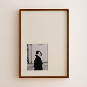 May include: A framed black and white photograph of a person in a black coat, looking over their shoulder. The frame is a dark brown color, and the background is a cream color.