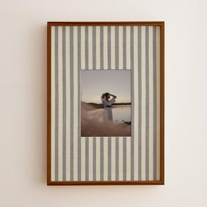 May include: A rectangular picture frame with a dark wood finish and a fabric mat featuring vertical gray and cream stripes. The frame contains a photograph of a person in a white garment standing on a beach at sunset near the water.