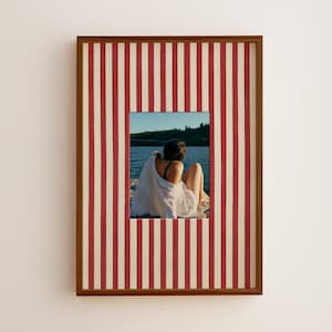 Pode incluir: Uma peça de arte de parede retangular com uma fotografia de uma pessoa de camisa branca sentada junto à água, emoldurada em madeira castanha com um passe-partout com um padrão de riscas verticais vermelhas e brancas à volta da imagem.