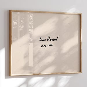 Puede incluir: Un cuadro enmarcado con la frase "how blessed are we" en escritura negra sobre un fondo beige. El marco es de madera clara. El cuadro se exhibe en una pared blanca.
