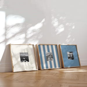 May include: Three framed photographs with wooden frames lean against a white wall. The frames contain black and white images. One frame has a white background, another has blue and white stripes, and the third has a solid blue background.