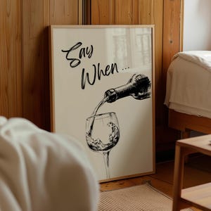 May include: Framed art print featuring a black and white illustration of wine being poured into a glass. The text "Say When..." is written in a cursive font at the top of the print. The artwork is displayed in a light wood frame.