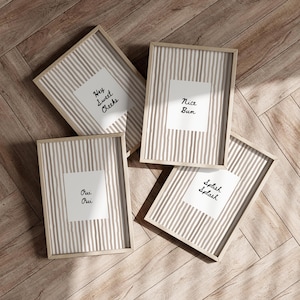 Op de afbeelding: Vier ingelijste kunstafdrukken met licht houten frames. Elke print heeft een witte achtergrond met een verticaal gestreept patroon in beige en wit. Elke print heeft een witte vierkant met een andere zin in zwart script: "Hey Sweet Cheeks", "Nice Bum", "Oui Oui" en "Splash Splash".