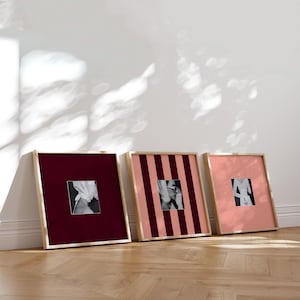 Può includere: Tre stampe artistiche quadrate incorniciate con cornici in legno. Le stampe presentano immagini in bianco e nero su sfondi bordeaux, a righe rosa e rosa tinta unita. Le cornici sono appoggiate a un muro bianco su un pavimento in legno.