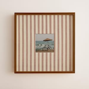 May include: A square wall art piece featuring a wooden frame and a mount with vertical dusty pink and cream stripes. The centre displays a photograph of a bicycle parked on a sandy beach under a straw umbrella with the ocean in the background.