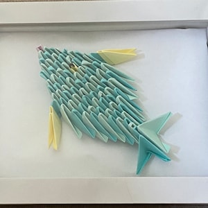 Pode incluir: Um peixe de origami emoldurado feito de papel azul e amarelo. O peixe é exibido em um fundo branco.