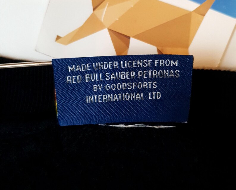 Puede incluir: Una etiqueta azul con texto blanco que dice "Made under license from Red Bull Sauber Petronas by Goodsports International Ltd."