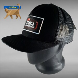 Könnte beinhalten: Schwarze Trucker-Kappe mit flachem Schirm und Mesh-Rückseite. Die Kappe hat einen rechteckigen Aufnäher mit dem Logo des Formel 1 Las Vegas Grand Prix in Weiß und Rot. Die Kappe wird auf einem schwarzen Schaufensterkopf präsentiert.