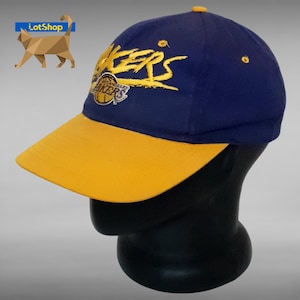 Puede incluir: Gorra de béisbol azul marino y amarilla con el logotipo de Los Angeles Lakers. La gorra tiene una visera amarilla y el nombre del equipo bordado en dorado. El logotipo de los Lakers también está bordado en la parte delantera.