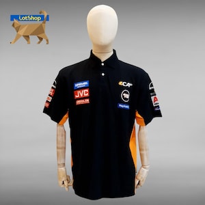 May include: Black polo shirt with orange side panels and various sponsor logos, such as Dremel, JVC, and PlayStation. The shirt has a button-down collar and short sleeves, presented on a mannequin.