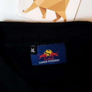 Puede incluir: Camiseta negra con un logotipo de Red Bull Sauber Petronas azul y rojo. La camiseta tiene una etiqueta de talla que dice "XL".