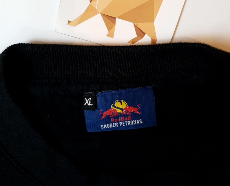 Puede incluir: Camiseta negra con un logotipo de Red Bull Sauber Petronas azul y rojo. La camiseta tiene una etiqueta de talla que dice "XL".