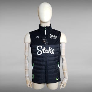 Peut inclure: Gilet matelassé noir et blanc avec le mot "Stake" en écriture blanche sur la poitrine. Le gilet a un col montant et une fermeture éclair. Le gilet porte les mots "Stake F1 Team" en écriture blanche. Le gilet a des accents verts.