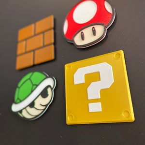 May include: Four colourful magnets featuring Super Mario Bros. characters. Includes a red and white mushroom, a green Koopa shell, a yellow question mark block, and a brown brick block. Ideal for decorating a fridge or any magnetic surface.