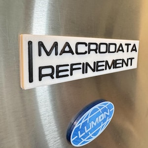 May include: A white rectangular magnet with the words "MACRODATA REFINEMENT" in black, bold letters. Below it is a blue oval magnet with the word "LUMON" in white, with a globe design. Both magnets are on a metallic surface.