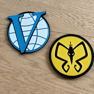 May include: Two round, black-edged patches. One features a blue "V" over a white globe. The other has a yellow background with a black butterfly-like design. Both are on a light wood surface.