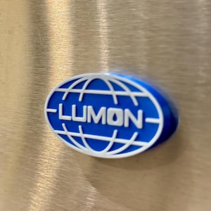 May include: A blue and white oval-shaped logo with the word "LUMON" in white lettering. The logo is set against a metallic, brushed metal background. The logo features a globe design.