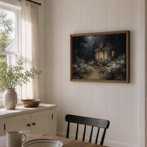May include: A framed landscape painting depicts a cottage illuminated by warm light, nestled in a forest with a path leading to it. The artwork is in a wooden frame, hanging on a white paneled wall. The room also features a wooden table, a vase with greenery, and a window.