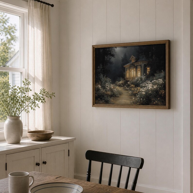 May include: A framed landscape painting depicts a cottage illuminated by warm light, nestled in a forest with a path leading to it. The artwork is in a wooden frame, hanging on a white paneled wall. The room also features a wooden table, a vase with greenery, and a window.