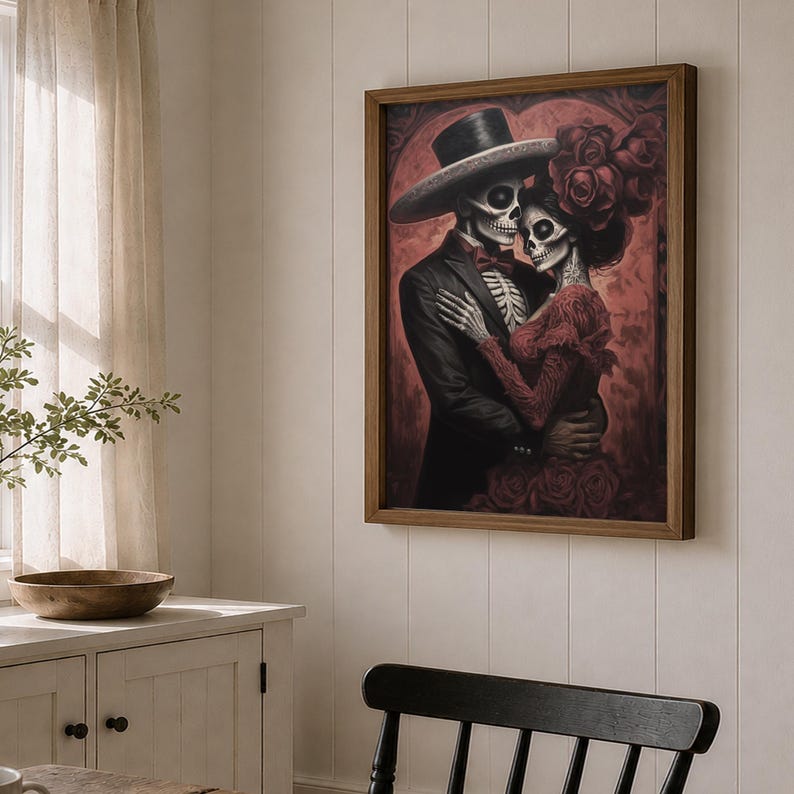 May include: A framed artwork shows a skeletal couple in an embrace. The man is in a black suit and top hat, the woman in a red dress and floral headpiece. The background is deep red, and the frame is a warm brown.