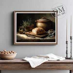 Op de afbeelding: Een ingelijste print van een stilleven met een rustieke keukensc&egrave;ne. Het kunstwerk toont een brood op een houten snijplank, een keramische pot, een klein glas vloeistof en een kruidenplant in een pot. De print is getiteld "Dark Moody Wall Art."