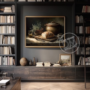 Op de afbeelding: Een ingelijst stilleven schilderij met een brood op een houten snijplank, een kleipot en een kruidenplant in een pot. Het schilderij heeft een warm kleurenpalet en wordt getoond op een donkergrijze muur. De tekst "Dark Moody Wall Art" is zichtbaar.