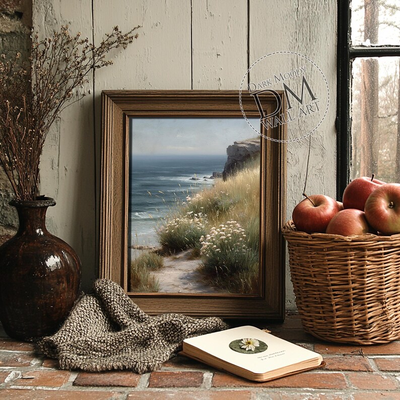 Puede incluir: Pintura enmarcada de un paisaje costero con un camino hacia el mar. La obra de arte est&aacute; en un marco de madera, mostrando un acantilado, hierba y flores blancas. Tambi&eacute;n se ve una cesta de manzanas rojas y un jarr&oacute;n.
