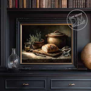 Op de afbeelding: Een ingelijst stilleven schilderij met een brood op een houten plank, een kleipot en een kleine glazen pot. Het kunstwerk heeft een donkere, melancholische esthetiek met warme tinten en een vintage uitstraling. Het schilderij is ingelijst in een zwart frame.