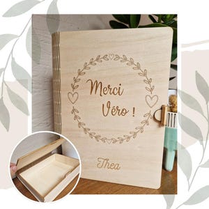 May include: A wooden box with a hinged lid. The box is engraved with the text "Merci Vero!" and "Thea" in a decorative font. The box is decorated with a wreath of leaves and hearts.
