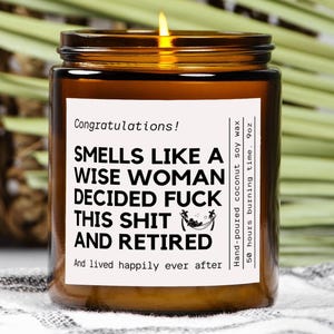 May include: A lit amber glass candle with a white label that reads "Smells like a wise woman decided fuck this shit and retired." The candle is 9 oz and made with hand-poured coconut soy wax. The candle has a 50-hour burn time.