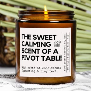 May include: A lit amber-colored candle in a glass jar with a white label. The label reads "THE SWEET CALMING SCENT OF A PIVOT TABLE" with additional text. The candle is 9oz and made with hand-poured coconut soy wax.