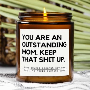 May include: A lit candle in a brown glass jar with a white label. The label reads "YOU ARE AN OUTSTANDING MOM. KEEP THAT SHIT UP." The candle is hand-poured coconut soy wax and has a 255g volume with a 45-hour burn time.