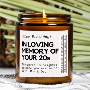 May include: A brown glass candle with a white label that reads "Happy Birthday! In Loving Memory of Your 20s The world is brighter because you are in it! Love, Mom & Dad" and "Hand-poured coconut soy wax 50 hours burning time. 9oz". The candle is sitting on a white and green surface. The candle is 9 ounces and is amber in color.