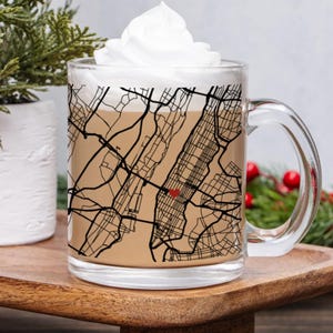 May include: A clear glass mug filled with a light brown beverage and topped with whipped cream. The mug features a black map design of a city, with a red heart marking a location. The mug sits on a wooden tray, with greenery in the background.