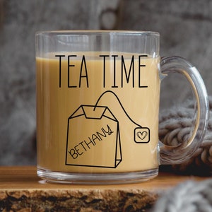 May include: A clear glass mug with a black outline of a tea bag that says "Bethany" and the text "TEA TIME". The mug is filled with a light brown liquid.