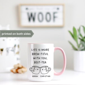 May include: White ceramic mug with a pink rim and handle. The mug has a black graphic that says "Life is more brew-tiful with you, best-tea" with two cartoon teacups with faces. The text "Sarah Christina" is below the teacups.