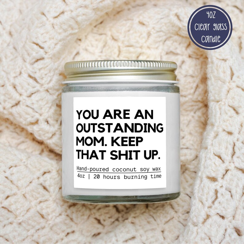 May include: A clear glass candle jar with a silver lid, featuring a white label with black text that reads "YOU ARE AN OUTSTANDING MOM. KEEP THAT SHIT UP." The label also states "Hand-poured coconut soy wax 113g | 20 hours burning time."