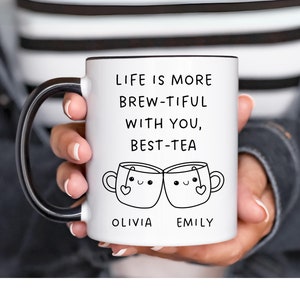 May include: White ceramic mug with a black rim and handle. The mug has a black and white illustration of two teacups with faces and the text "Life is more brew-tiful with you, best-tea Olivia Emily".
