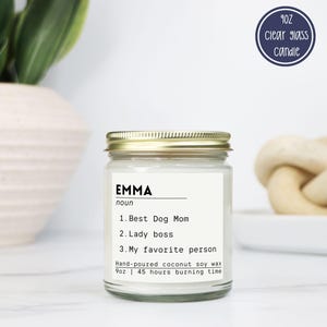 May include: A 9oz clear glass candle with a gold lid and a white label featuring the text: EMMA noun 1. Best Dog Mom 2. Lady boss 3. My favorite person. The label also states: Hand-poured coconut soy wax 9oz | 45 hours burning time.