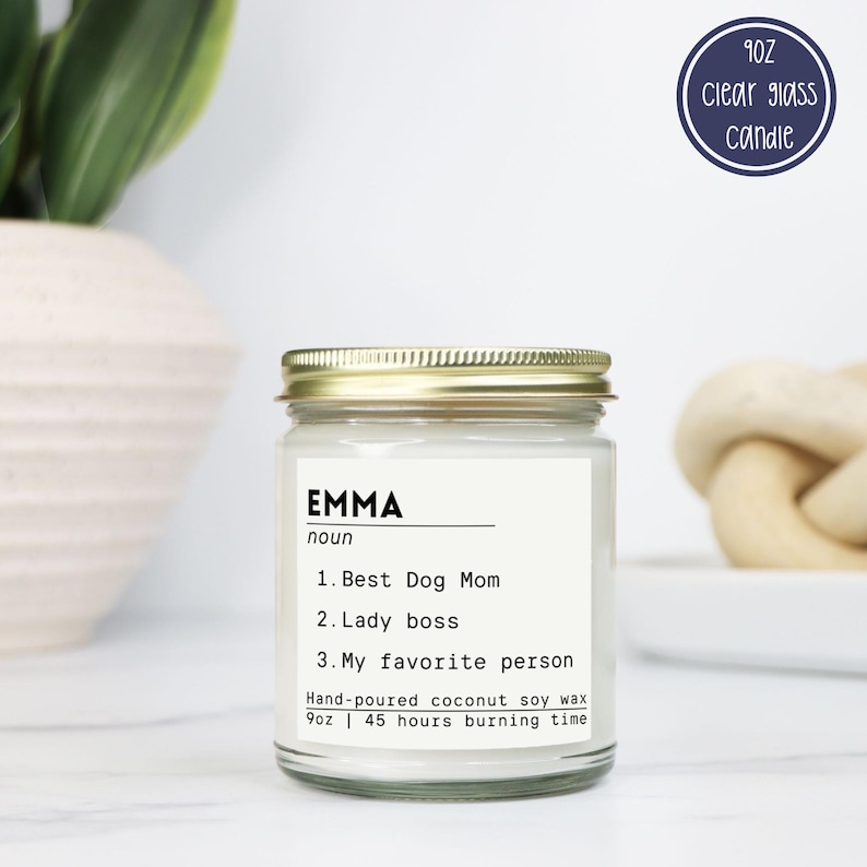 May include: A 9oz clear glass candle with a gold lid and a white label featuring the text: EMMA noun 1. Best Dog Mom 2. Lady boss 3. My favorite person. The label also states: Hand-poured coconut soy wax 9oz | 45 hours burning time.