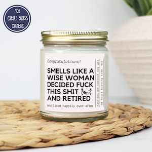 May include: A clear glass candle jar with a gold lid. The label reads "Smells Like a Wise Woman Decided Fuck This Shit and Retired." The candle is hand-poured coconut soy wax and has a 50-hour burn time. A blue circle indicates the candle is 9oz.