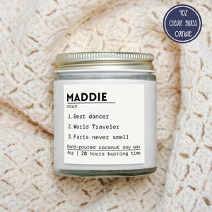 May include: A 4oz clear glass candle with a silver lid and a white label. The label features the text MADDIE noun, 1. Best dancer, 2. World Traveler, 3. Farts never smell, Hand-poured coconut soy wax, 4oz | 20 hours burning time.