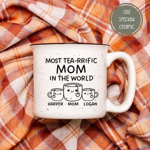 Może przedstawiać: Biały kubek ceramiczny z czarną obw&oacute;dką i uchwytem. Na nim napis "Most tea-rrific Mom in the world" z trzema kresk&oacute;wkowymi filiżankami do herbaty pod spodem. Filiżanki do herbaty są oznaczone jako "Harper", "Mom" i "Logan".