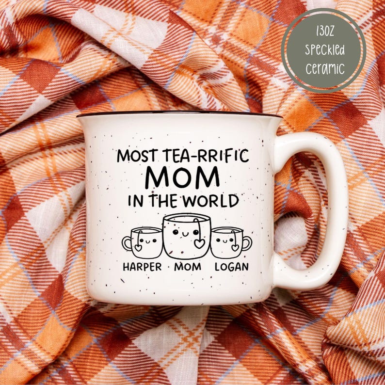 Może przedstawiać: Biały kubek ceramiczny z czarną obw&oacute;dką i uchwytem. Na nim napis "Most tea-rrific Mom in the world" z trzema kresk&oacute;wkowymi filiżankami do herbaty pod spodem. Filiżanki do herbaty są oznaczone jako "Harper", "Mom" i "Logan".
