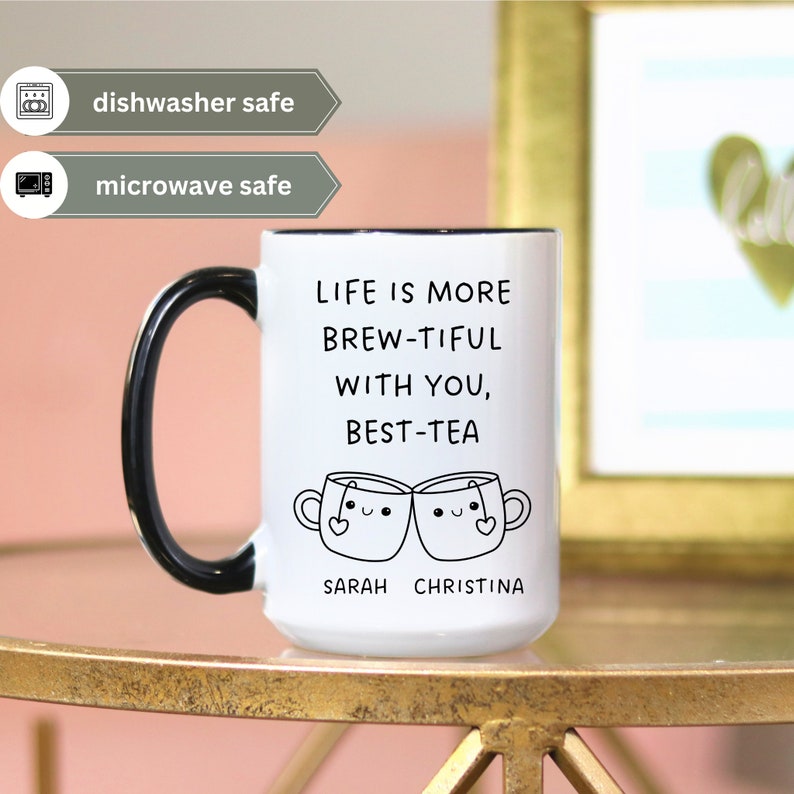 May include: White ceramic mug with a black handle and rim. The mug has a black and white illustration of two teacups with faces and the text "Life is more brew-tiful with you, best-tea" and the names "Sarah Christina".